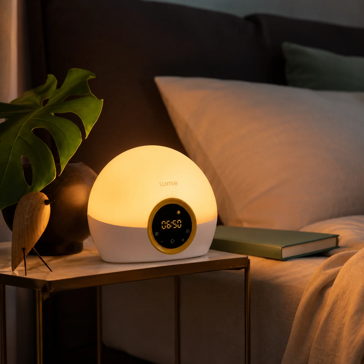 Lumie Bodyclock Rise100 alarm clock glowing warmly on a bedside table next to a bed, plant, and book in a cozy evening bedroom scene.